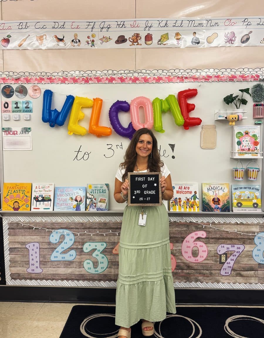 Jennifer Klingerman in her classroom on the first day of the school year, holding a sign that reads first day of 3rd grade, under a Welcome to 3rd grade display